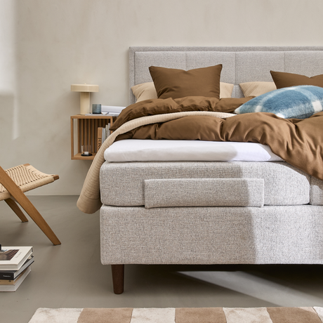 Ljust sovrum med stoppad säng i grå textil, brunt bäddset och kuddar, blårutad kudde, väggmonterad trähylla med lampa och böcker, flätad stol och beige-vitrutig matta.