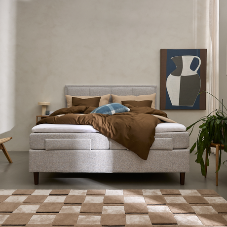 Grå kontinentalsäng med bruna sängkläder, blå prydnadskudde, tavla med vas, grön växt och beige‑brun rutmönstrad matta i ett ljust sovrum.