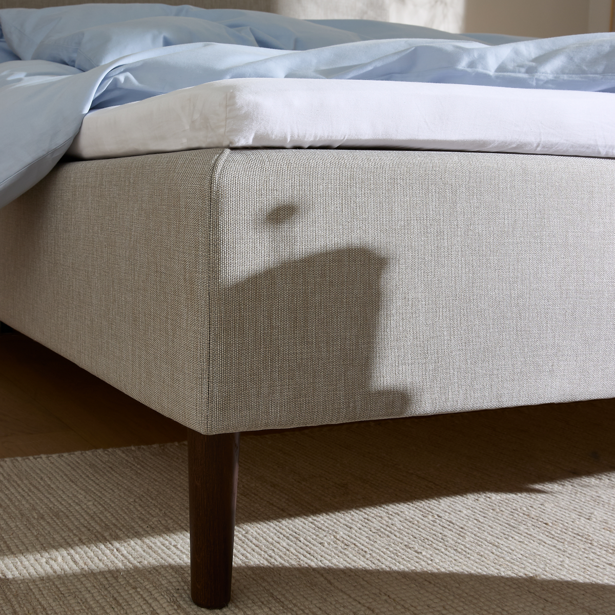 Närbild av sänghörn med beige klädd sängram, vit bäddmadrass och ljusblått täcke; mörkt träben på matta i solsken.