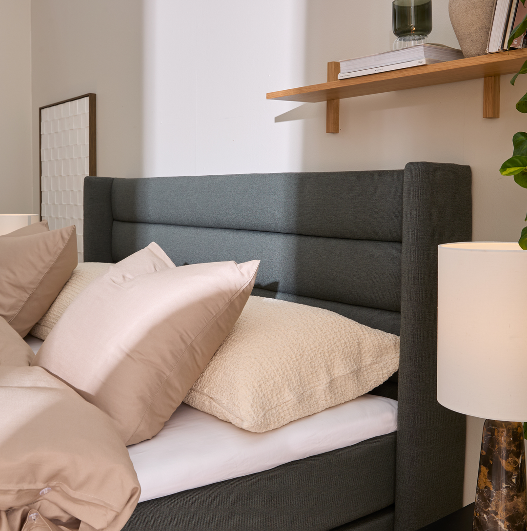 Närbild på säng med grå stoppad gav, beige kuddar och bordslampa; trähylla med böcker och vas i mjukt dagsljus.