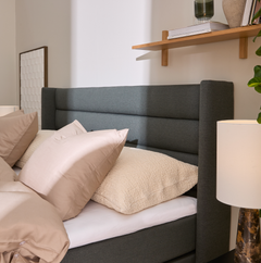 Närbild på säng med grå stoppad gav, beige kuddar och bordslampa; trähylla med böcker och vas i mjukt dagsljus.