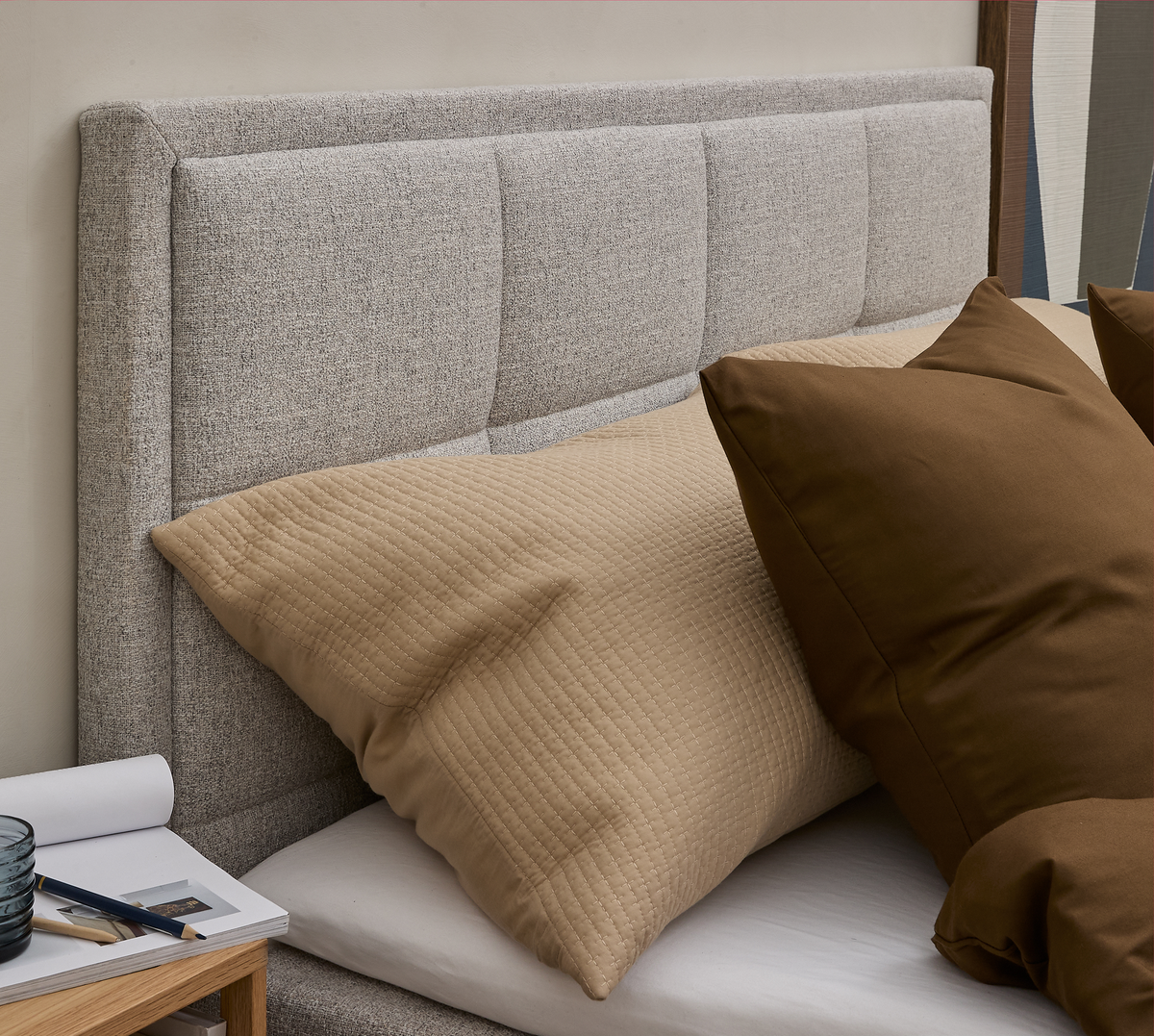 Stoppad grå sänggavel med en beige quiltad kudde och en brun kudde på sängen, bredvid ett litet nattduksbord med glas och pennor.