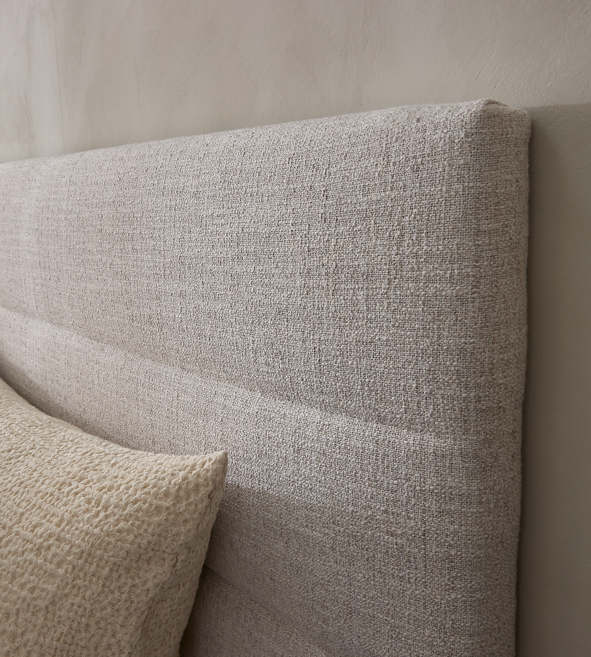 Närbild av en beige textilklädd sänggavel med en strukturerad kudde mot en neutral vägg.