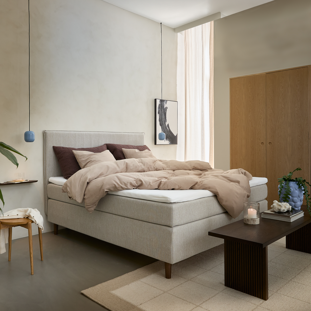 Ljust, minimalistiskt sovrum med beige stoppad kontinentalsäng bäddad med sandfärgat täcke och bruna kuddar, två blå pendellampor, mörkt soffbord med tändt ljus och böcker, ljus matta och garderob i trä.