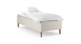 Enkelbädd med beige klädd sängbas, träben och vit bäddning med kudde._ps