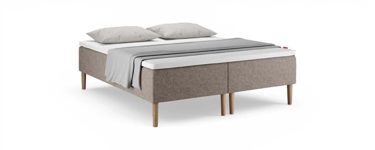 Kontinentalsäng i beige textil med träben, vit bäddmadrass, två kuddar och grå pläd på vit bakgrund.