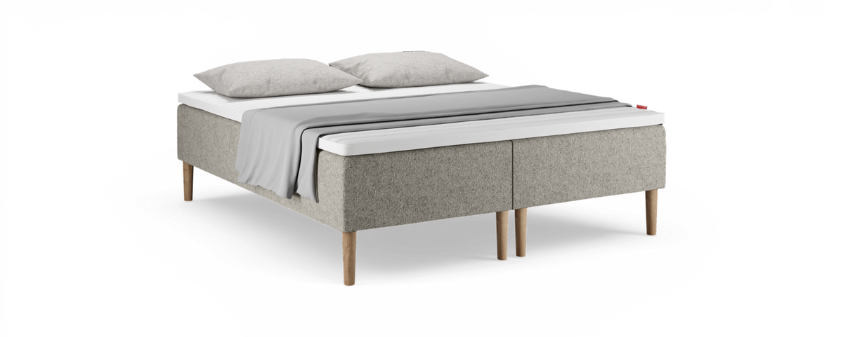 Beige klädd säng med träben, vit bäddmadrass, två grå kuddar och en ljusgrå pläd över bädden.