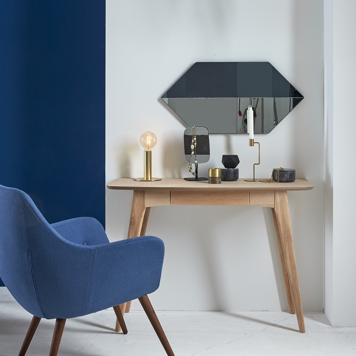 Ljusgrå vägg med blå accent, en träkonsol med geometrisk väggspegel ovanför, mässingsbordslampa med glödlampa, ljusstake och små dekorationer; blå fåtölj i förgrunden.