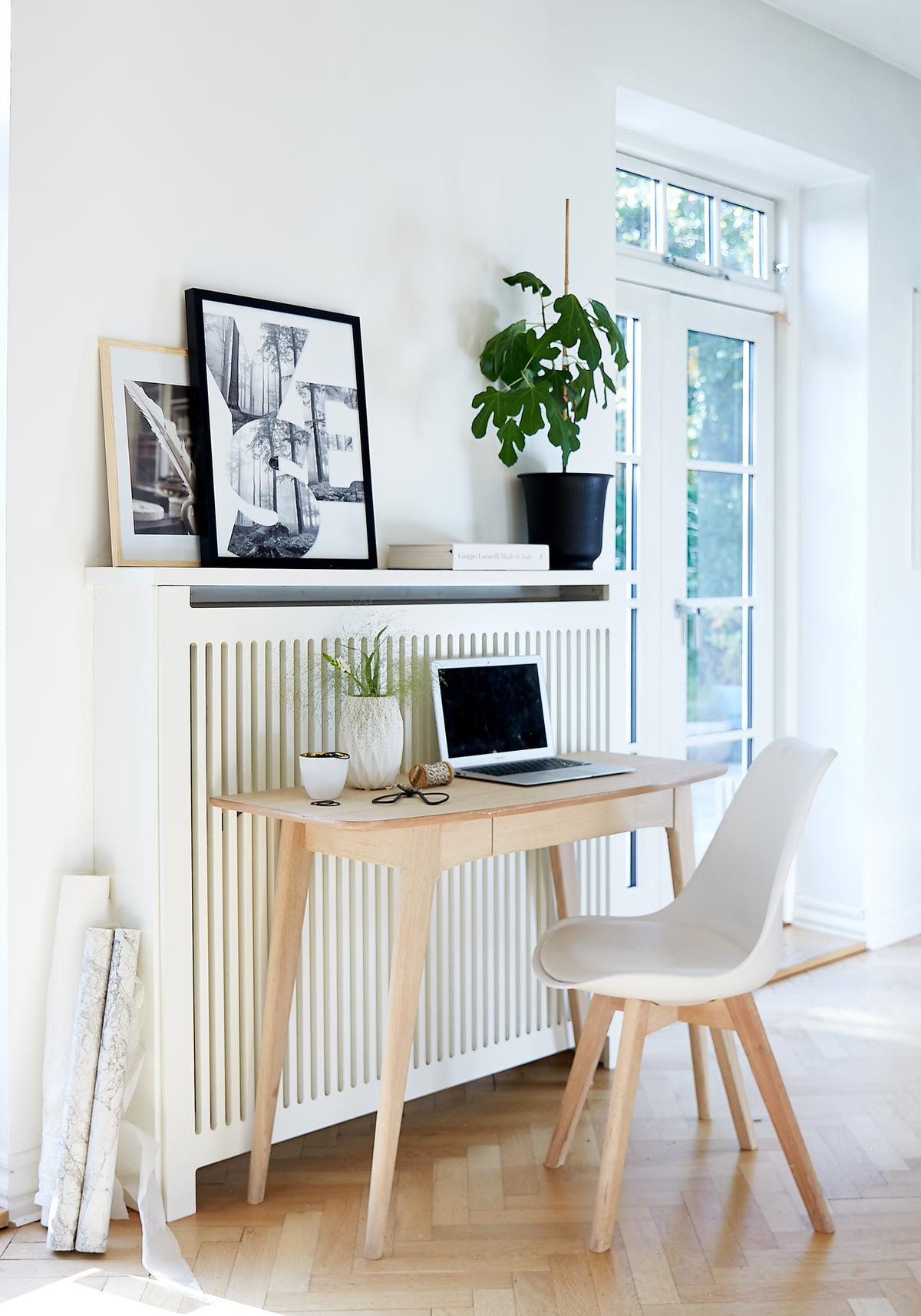 Ljus skandinavisk arbetsplats med ljust träbord, vit stol, laptop, grön krukväxt och YES‑konst framför fönster i dagsljus.