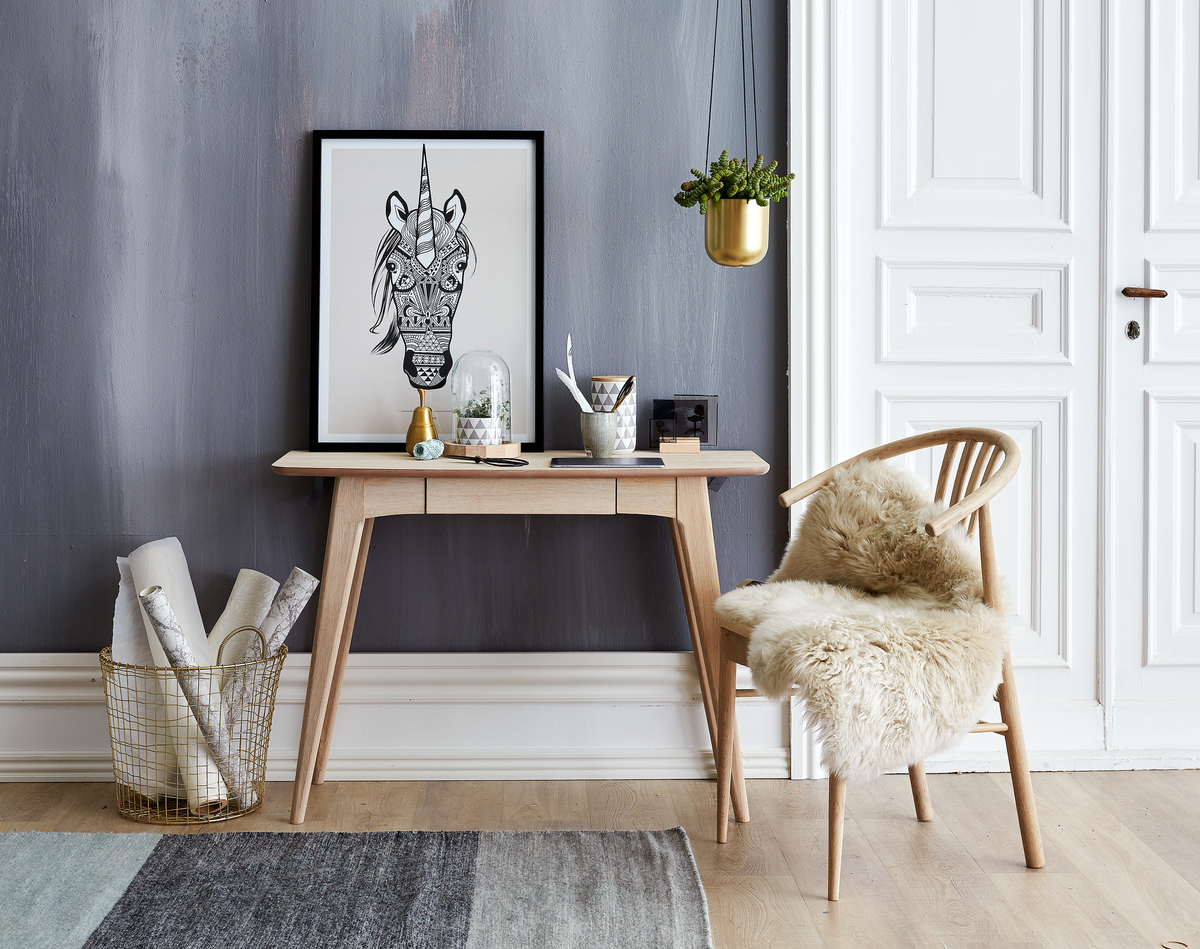 Ljus skandinavisk entré med konsolbord i ljust trä, stol med fårskinn, hängande guldfärgad kruka och en inramad svartvit illustration av en enhörning.