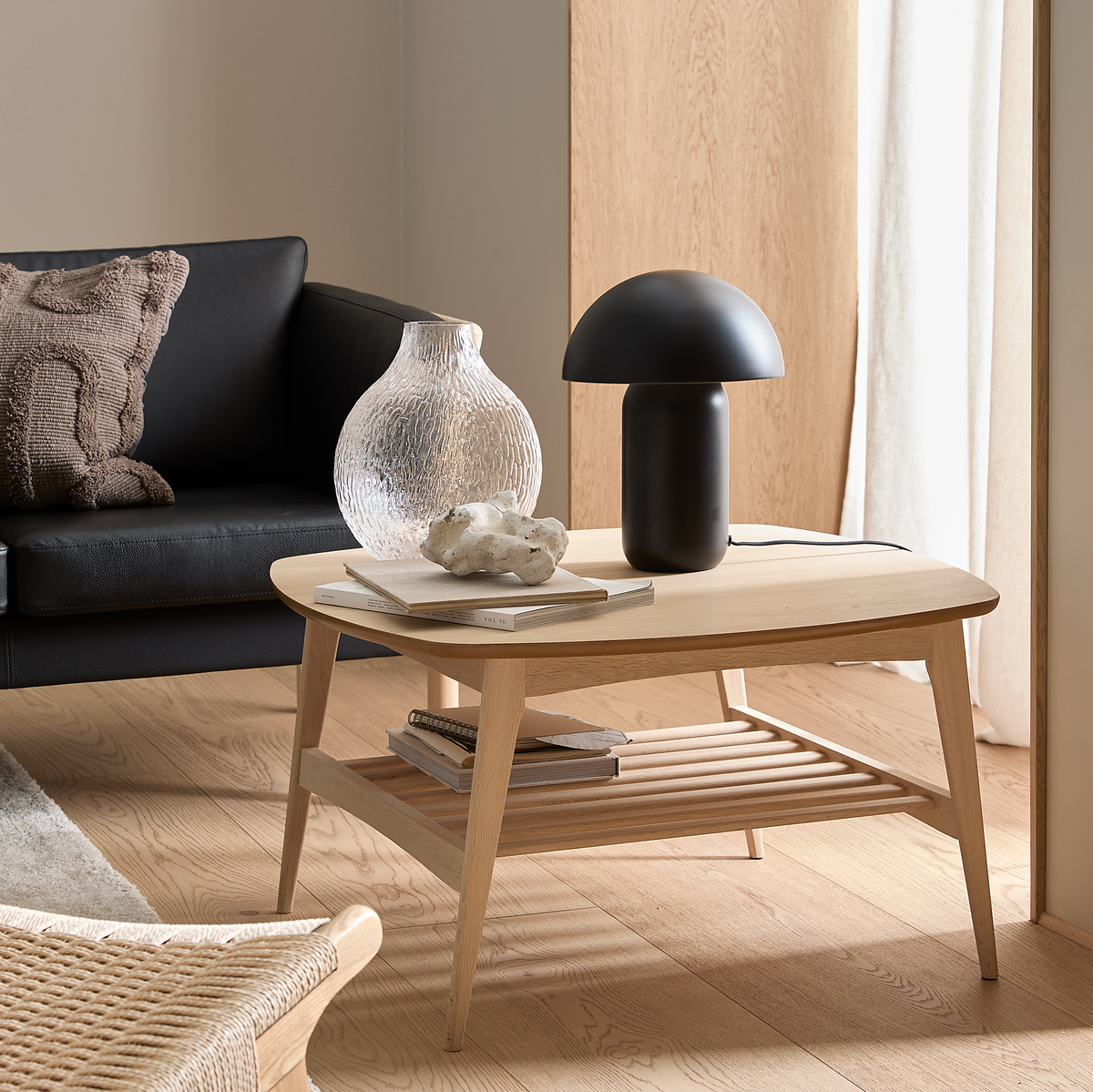 Skandinavisk vardagsrumsvy med ljus ek-soffbord med svart svamplampa, strukturerad glasvas, böcker och sten; svart lädersoffa, vävd stol och ljusa gardiner på ekgolv i mjukt dagsljus.