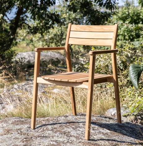 Trästol med armstöd i teak placerad utomhus på en klippa i soligt, grönt landskap.