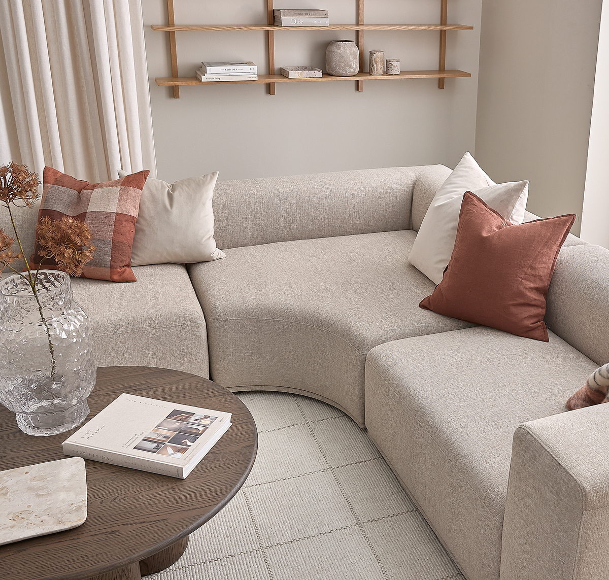 Ljusbeige modulsoffa med mjuka former och kuddar i terrakotta och vitt, intill ett runt träbord med boken ”Soft Minimal”, en ljus stenplatta och en klar strukturerad vas med torkade kvistar; i bakgrunden ljusa gardiner och enkla hyllor med böcker och keramik.