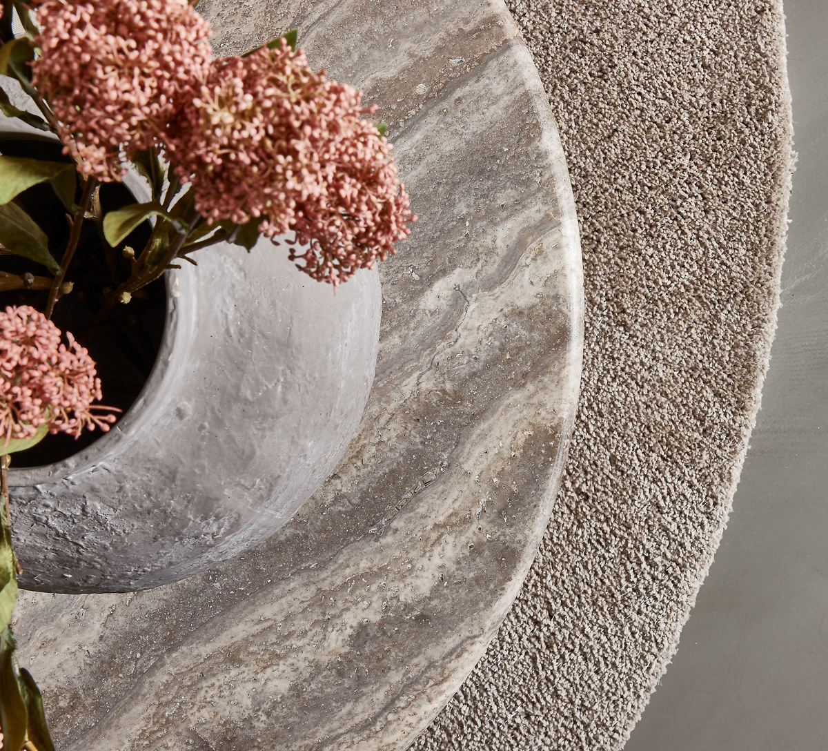 Närbild av ett runt stenbord med grå vas och rosa blommor på en beige, rund texturerad matta.