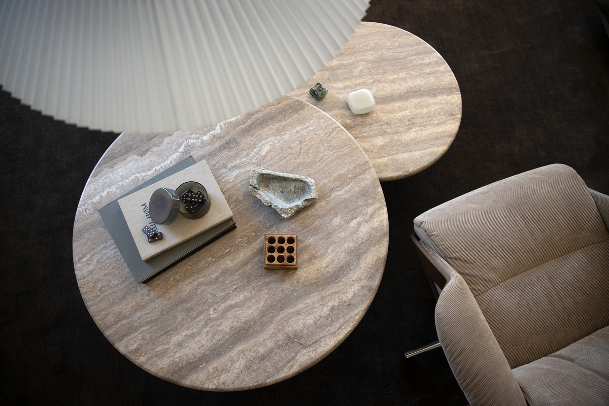 Två runda soffbord i beige sten sedda ovanifrån, dekorerade med böcker, ett glasobjekt med dominobrickor, en rå stenskål, ett litet trägaller och små objekt; bredvid syns en beige fåtölj och en plisserad taklampa.