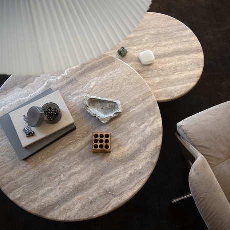 Runda soffbord i travertin med böcker (NOW), ett litet glas med dominobrickor, en dekorativ sten och en liten träbricka; beige fåtölj bredvid.
